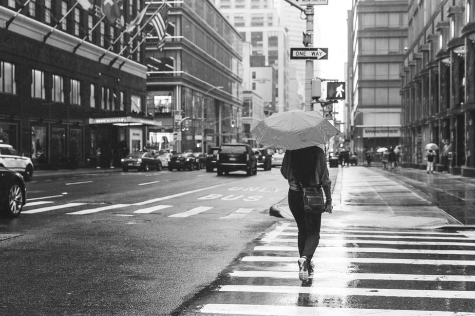 a person with an umbrella walking across a zebra crossing on a rainy day in a city setting. The black and white photo captures the rainy atmosphere, with buildings and vehicles in the background, including cars and what appears to be a bus. Street signs, such as “ONE WAY,” are also visible, contributing to the urban feel of the scene.